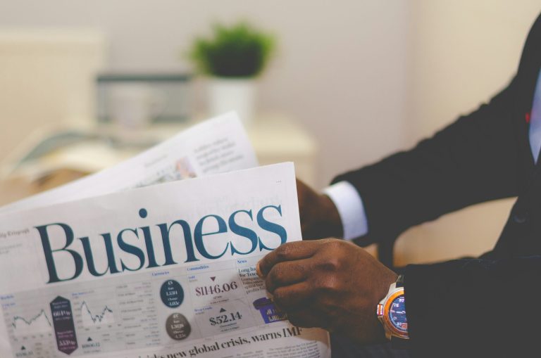 A person in a business suit holding a newspaper with the headline "Business" and financial data charts visible